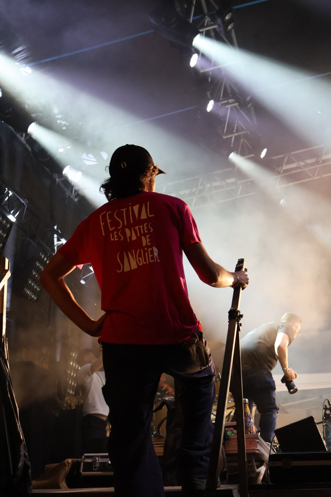 Sur scène au Festival des Pattes de Sanglier : un membre de l'équipe de dos, en t-shirt du festival, sous les faisceaux des projecteurs